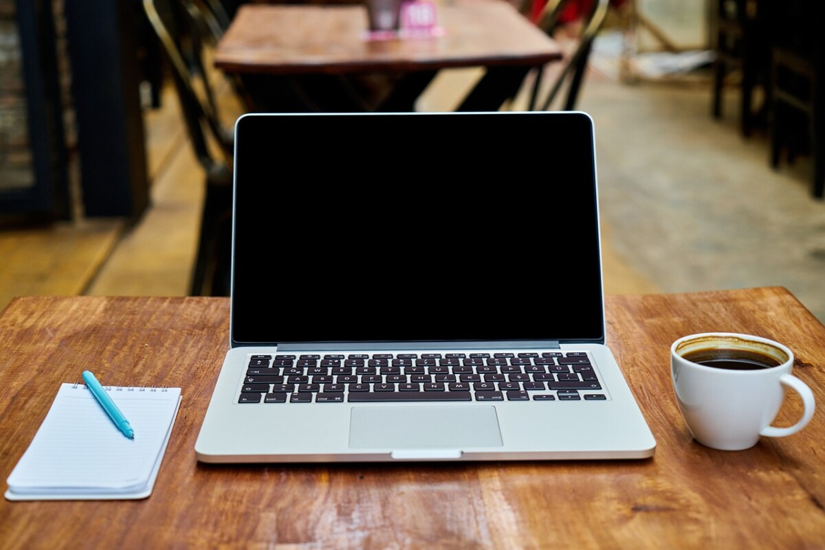 Auf einem braunen Tisch sind link ein Block mit Stift, in der Mitte ein aufgeklappter Laptop und rechts eine Kaffeetasse zu sehen
