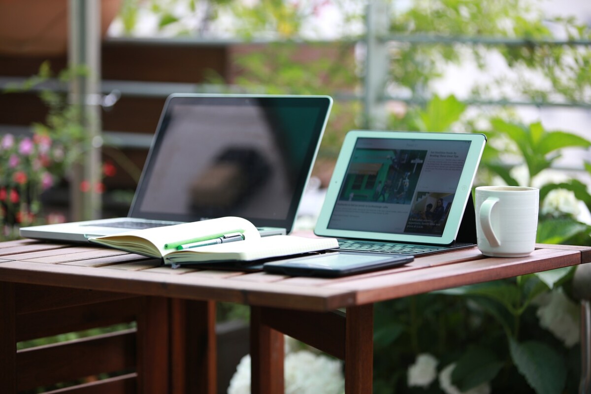 Ein Tisch auf dem ein Laptop und ein Tablet zusehen sind, davor liegt ein aufgeklappter Notizblock mit einem darauf liegenden Stift