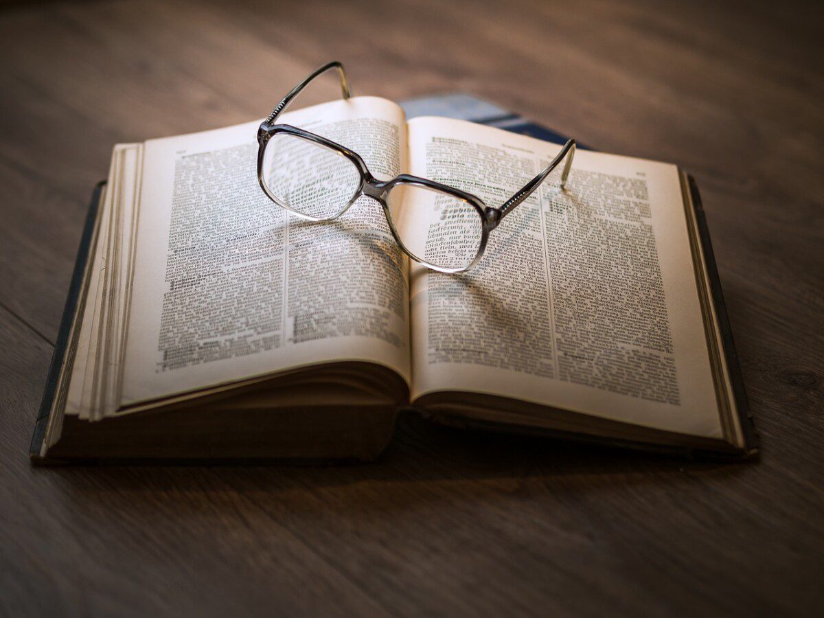 Das Foto zeigt ein aufgeschlagenes Buch, welches auf einem Parkettboden liegt. Auf dem Buch liegt eine Brille. Das Foto steht für die Literatursammlung bei einer Forschungsarbeit.