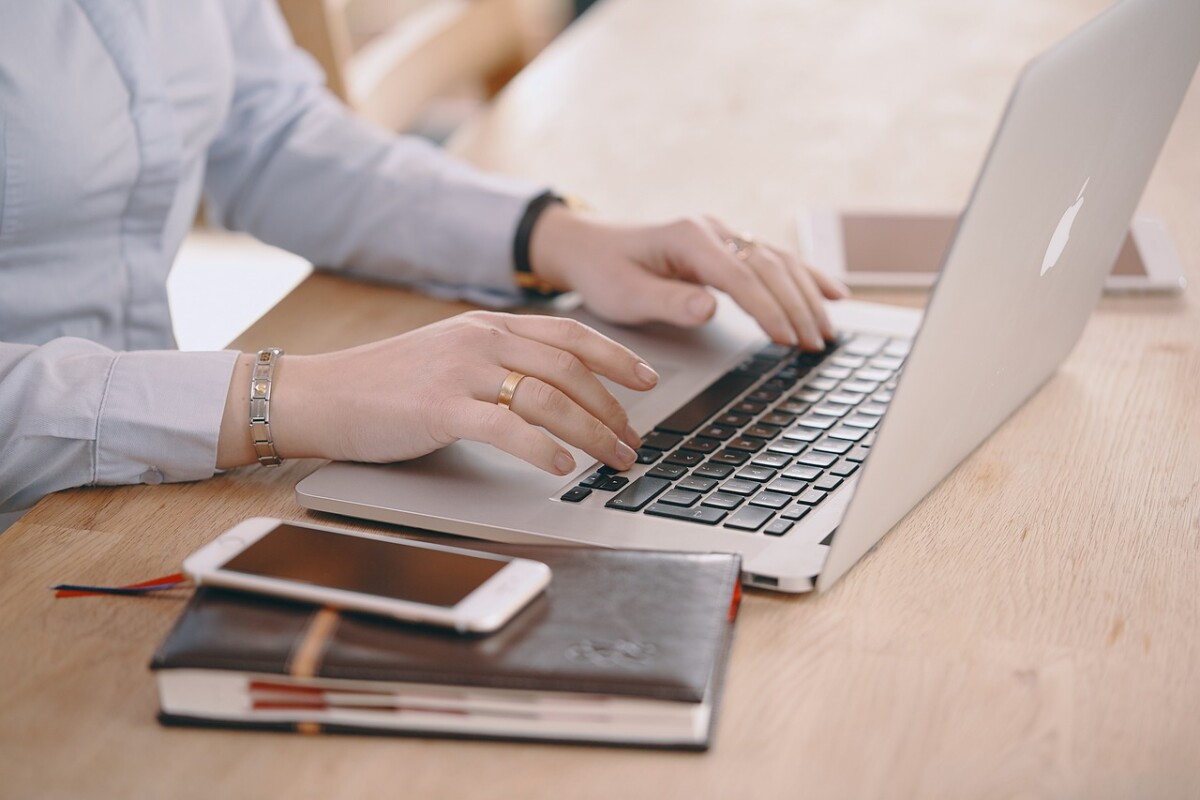 Redaktionelle Texte auf DeinText.at buchen. Auf dem Foto sieht man Hände einer weiblichen Person, die auf einem Laptop arbeitet. Neben dem Laptop liegen ein Notizblock, auf dem ein Handy positioniert ist. Der Laptop ist weiß und steht auf einem braunen Tisch.