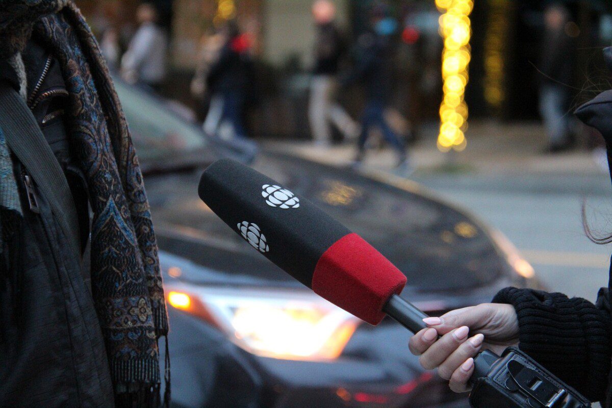 Das Foto zeigt eine Hand, die vor einem verschwommenen Hintergrund, der ein Auto zeigt, ein Mikrofon hält. Das Mikrophone zeigt auf eine dunkel gekleidete Person, bei der man nur den Torso sieht.