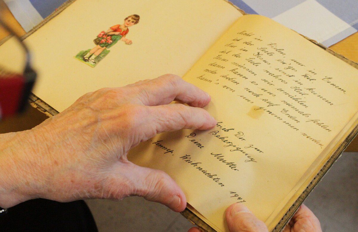 Auf dem Foto sieht man die Hände einer älteren Person, die gerade ein Buch in der Hand aufgeschlagen hält. Das Buch sieht nach einem Tagebuch aus.
